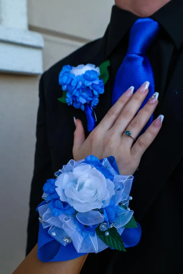 Blue rose corsage on a wrist beside a black suit with a matching boutonniere and blue tie before prom