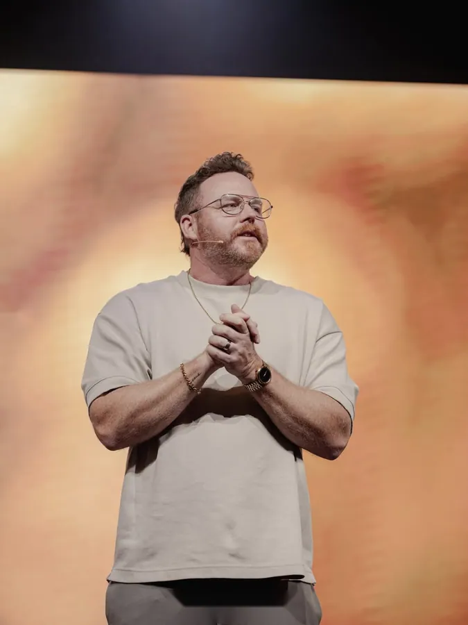 Man with glasses standing on stage with his hands clasped, speaking to the congregation
