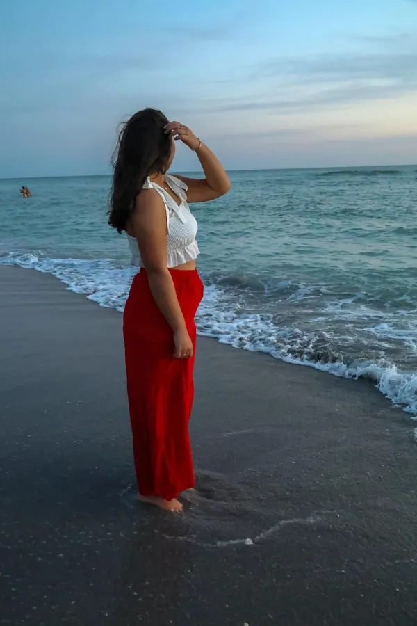 Senior in a white top and long red skirt standing barefoot in the shallow surf, looking out to the horizon