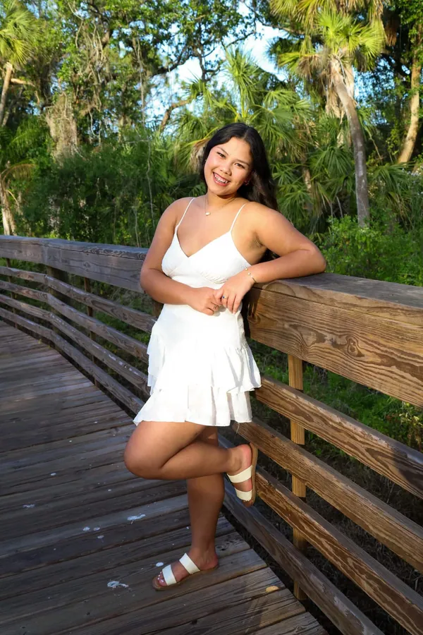 Woman in a white dress leaning against a wooden boardwalk railing surrounded by green foliage