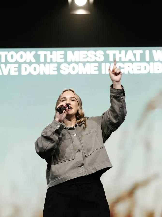Woman in a grey jacket singing on stage with one hand raised toward the light