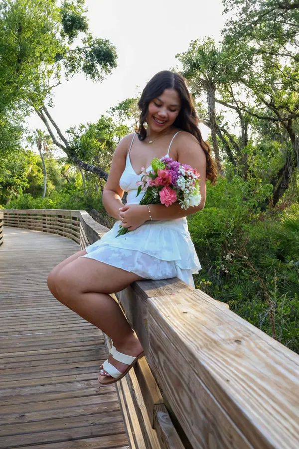 Woman in a white sundress sitting on a wooden boardwalk railing holding a bright floral bouquet
