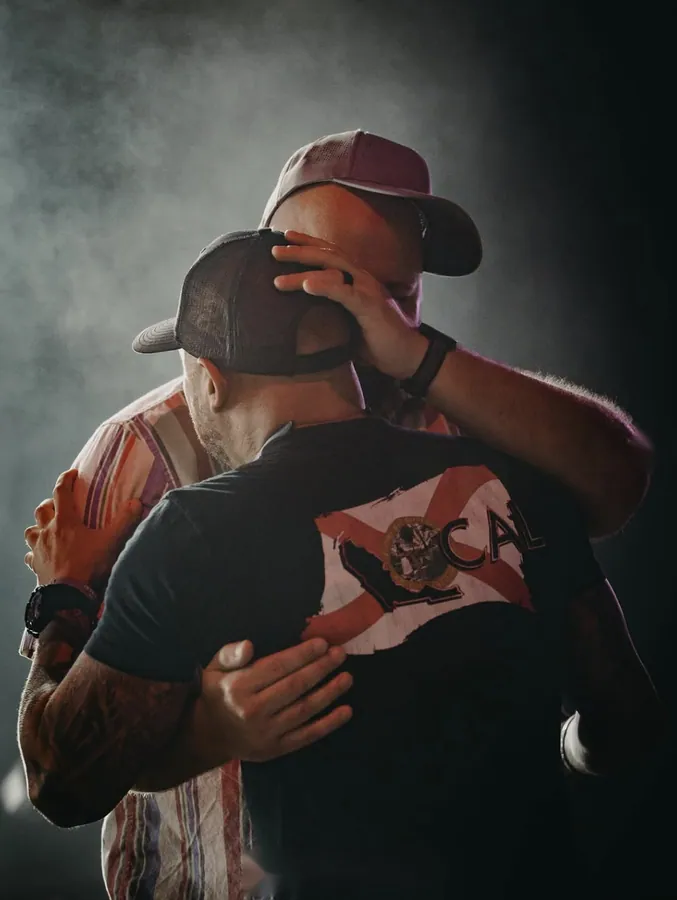 Two men in baseball caps embracing on stage under dramatic lighting