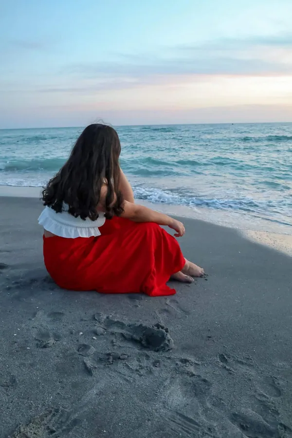 Senior in a red skirt sitting on the sand looking out at the Gulf as the light fades