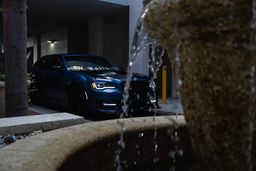 Deep teal Chrysler 300 parked in a shadowed brick courtyard, framed by the spray of a stone fountain at night
