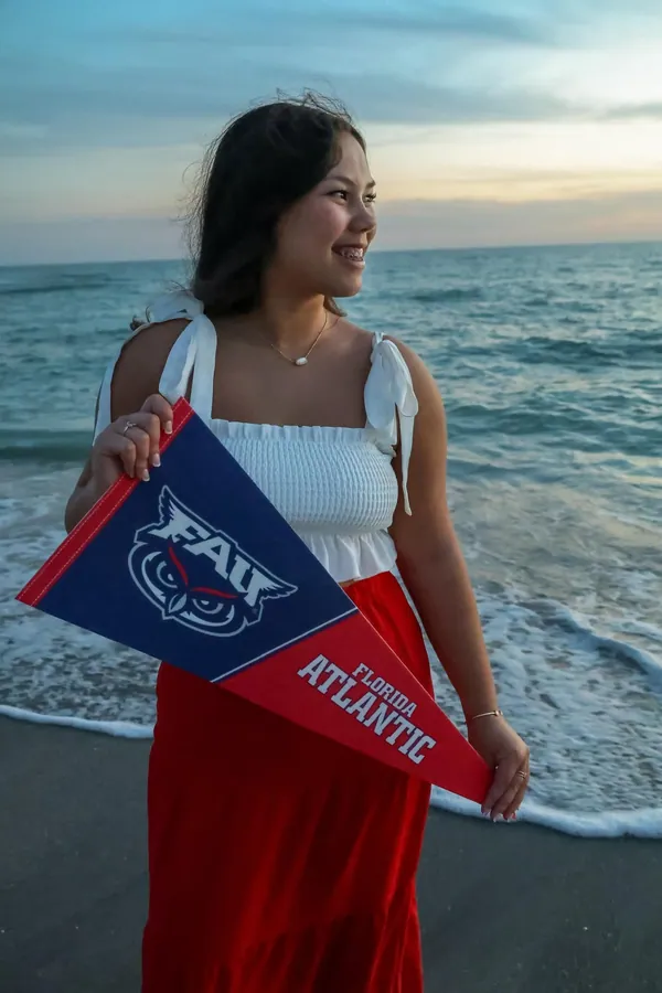 High school senior holding a Florida Atlantic pennant as waves wash over her feet at the shoreline