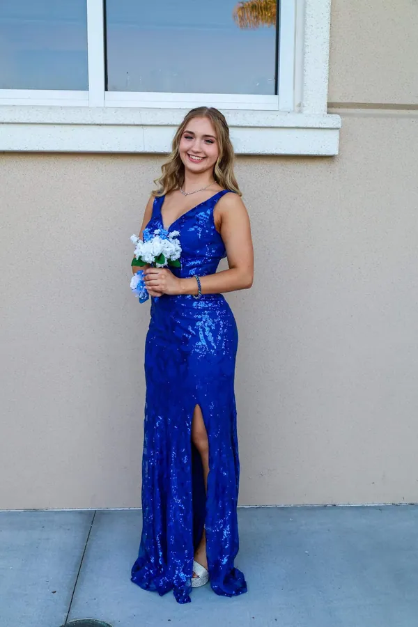 Young woman in a royal blue sequined prom dress standing against a warm tan wall holding a bouquet
