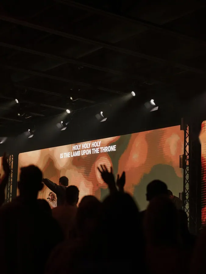 View from the back of a congregation toward a stage screen reading Holy Holy Holy Is The Lamb Upon The Throne