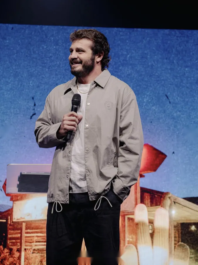Bearded man in a light jacket speaking into a microphone on stage