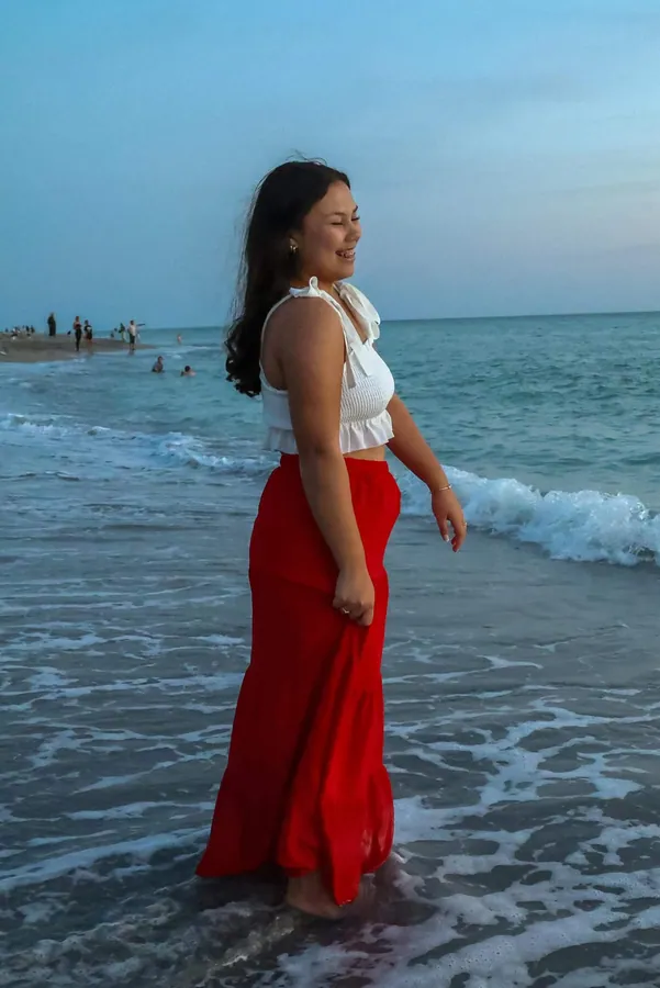 Senior in a white top and long red skirt walking through the ocean surf at twilight