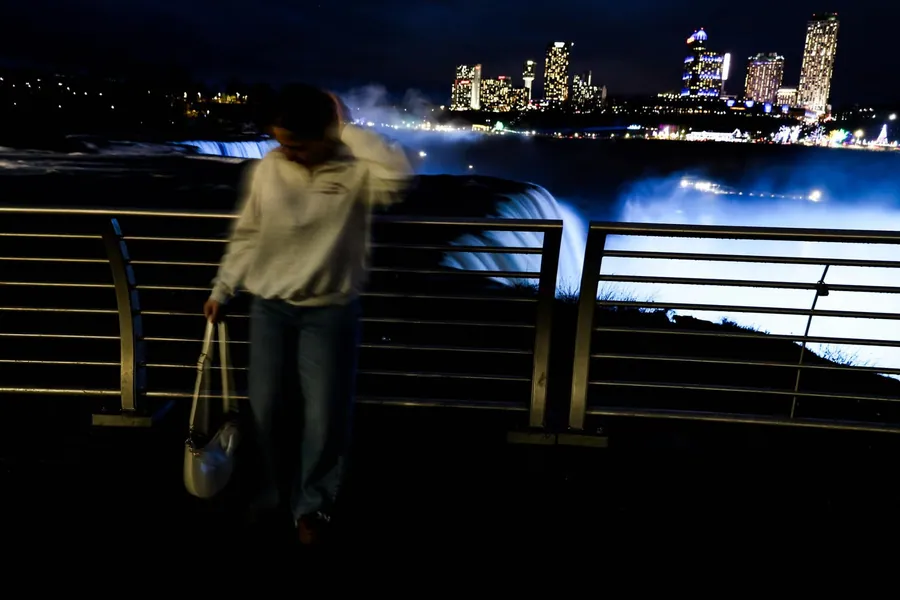 Long-exposure night portrait of a figure walking past a railing with illuminated waterfalls behind